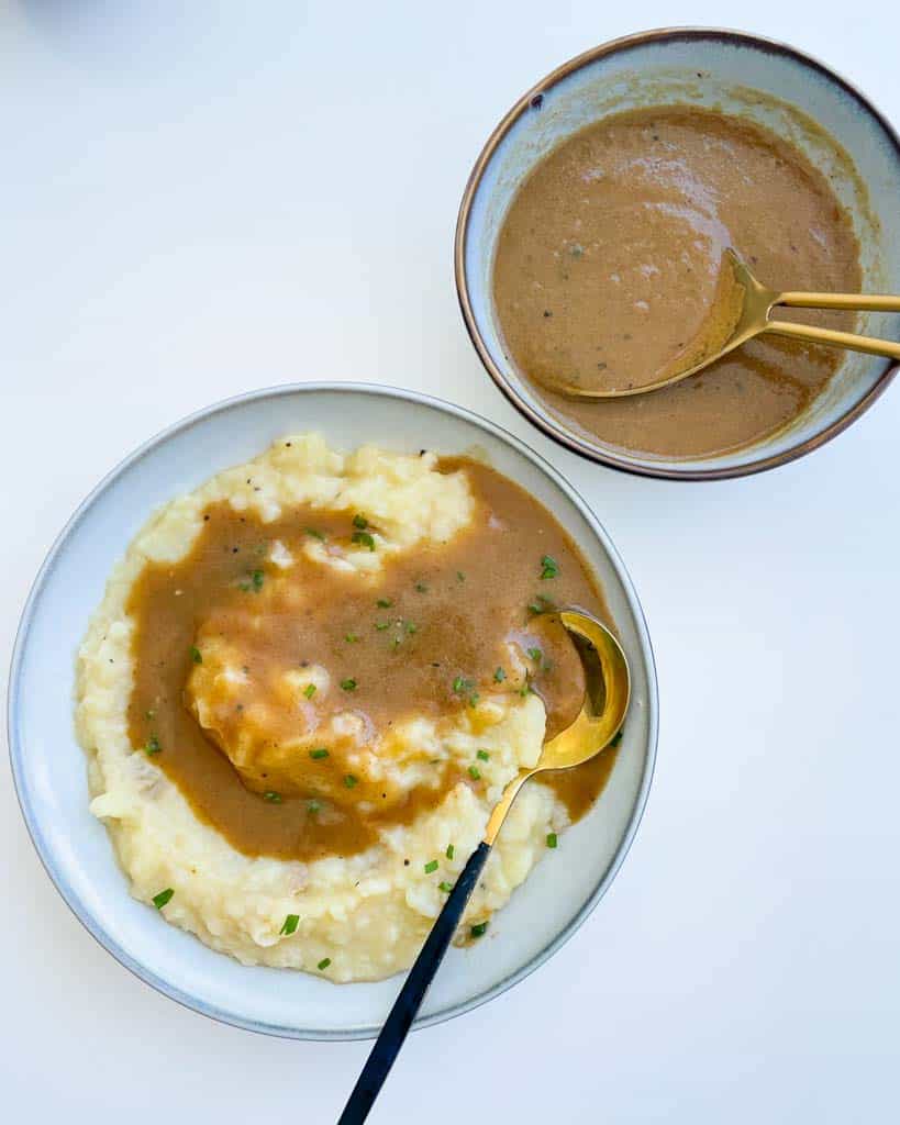 gluten free gravy on mashed potatoes in a white bowl with a gold spoon in the bowl and a bowl of gravy on the side with a large gold spoon in it