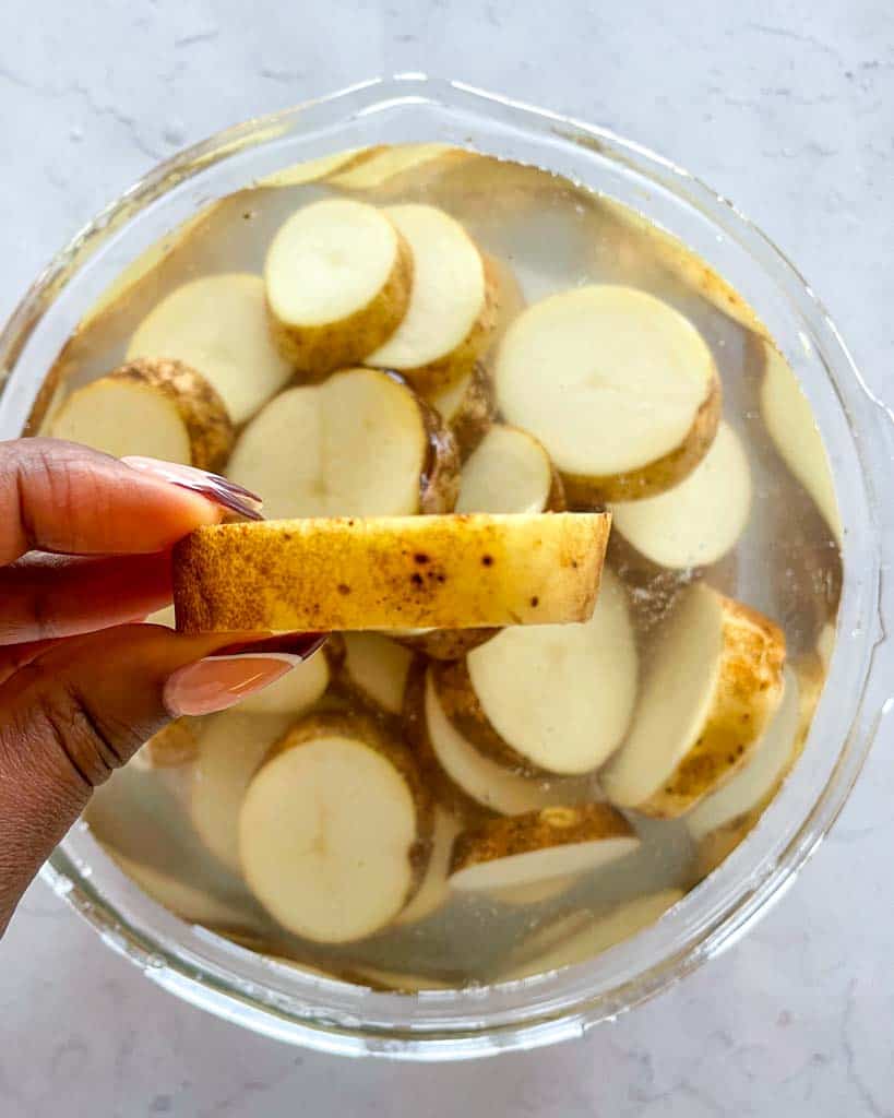 a bowl of sliced potatoes submerged in water with a hand holding one slice showing the thickness of the potato slice