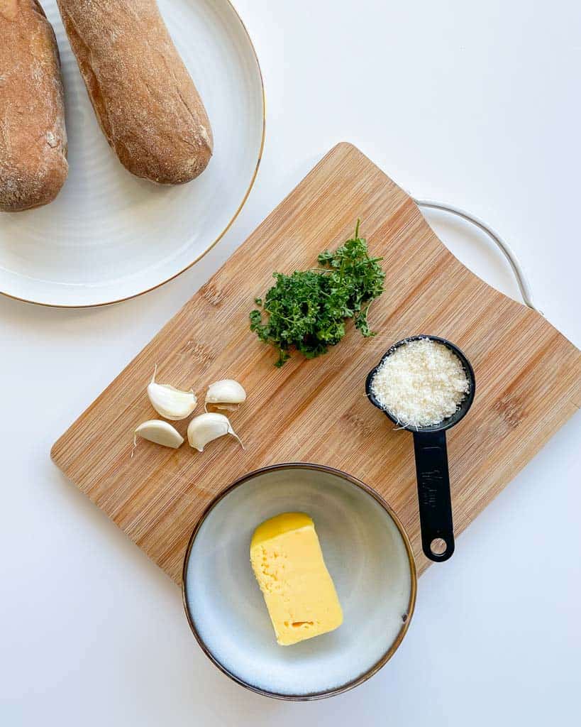 ingredients needed to make gluten free garlic bread. show are 2 loaves 4 garlic cloves fresh parsley a small bowl with butter a measuring cup with grated parmesan cheese