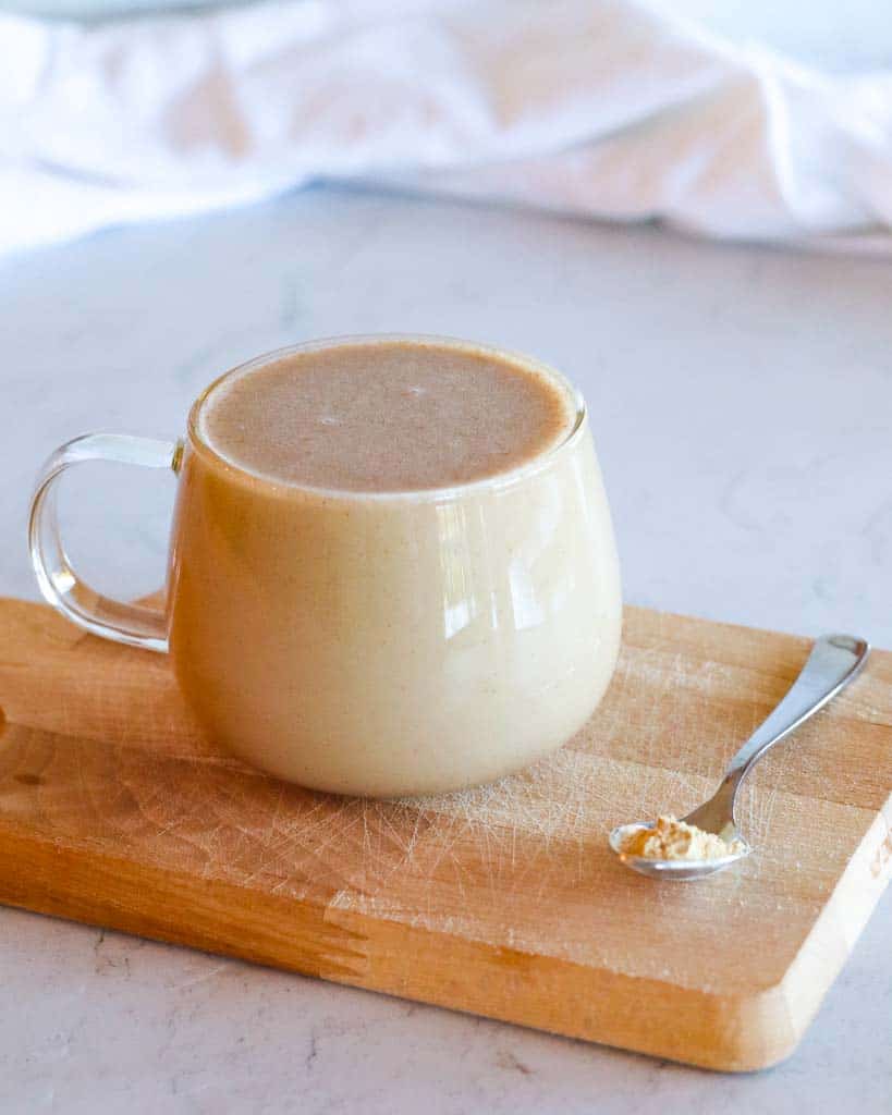 clear mug of maca coffee on a cutting board with a small spoon of maca powder on the side of the mug