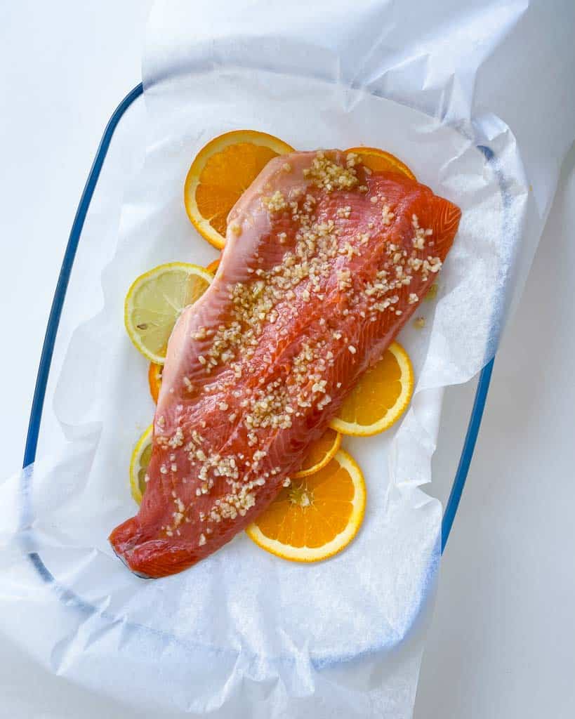 rainbow trout topped with minced garlic on sliced lemon and oranges in a parchment lined baking dish