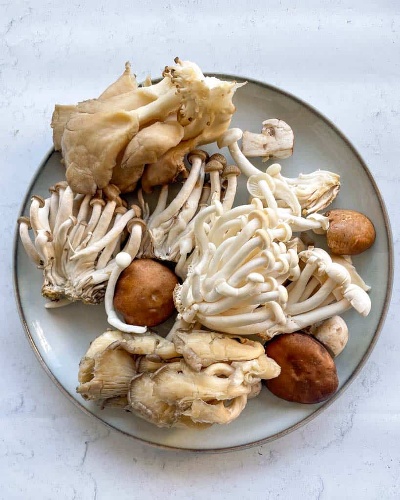 a white plate filled with a variety of mushrooms: shiitake mushrooms oyster mushrooms enoki mushrooms