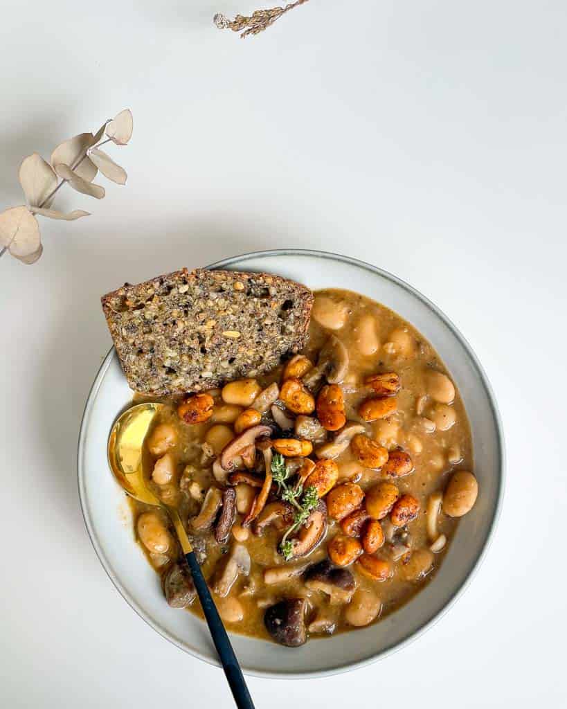 white bowl filled with white bean and mushroom soup topped with crispy white beans and fresh thyme with a piece of gluten free bread in the bowl and a gold and black spoon