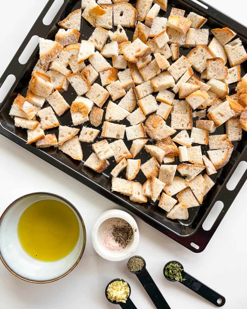ingredients to make gluten free croutons shown is gluten free bread cut into cubes on a sheet tray a small bowl of olive oil salt and pepper in a small ramekin and tablespoon of minced garlic and a half tablespoon of chopped rosemary and a teaspoon of dried herbs