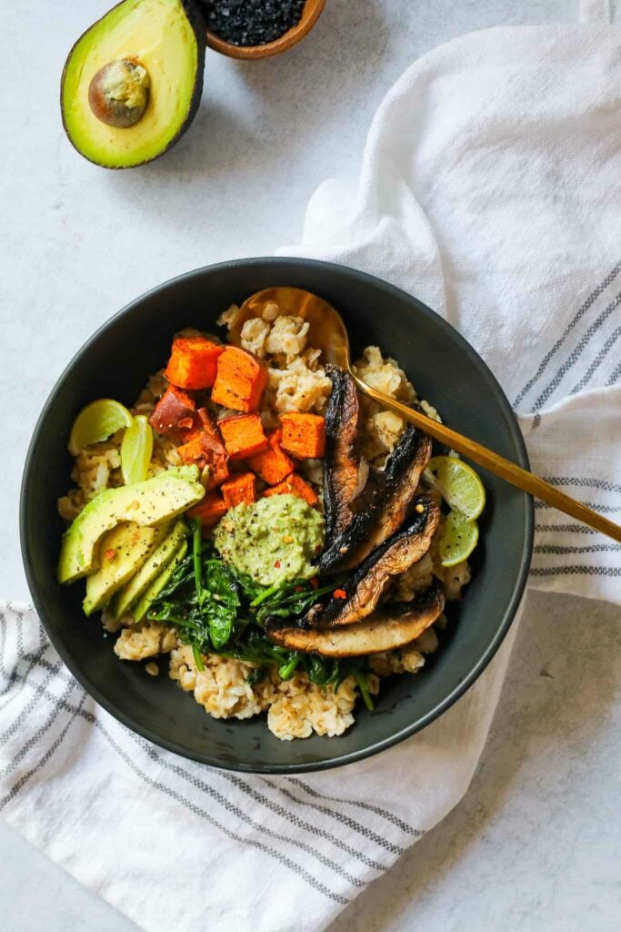 black bowl filled with savory oatmeal on the base and topped with sliced avocado, cooked mushrooms, roasted sweet potatoes, sauteed spinach, and pesto with a gold spoon in the bowl 