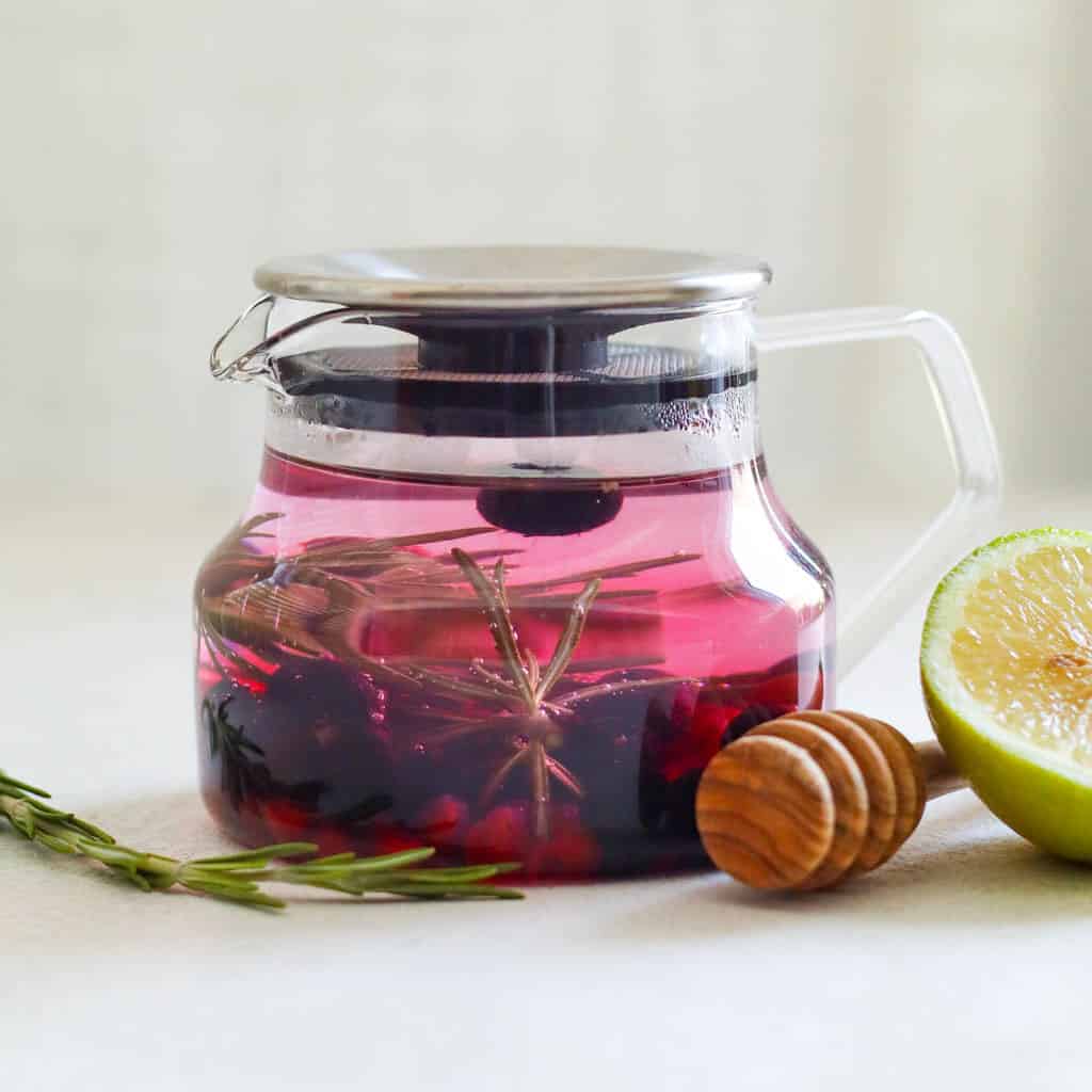 A glass tea infuser filled with blueberries, pomegranate, and rosemary to make rosemary tea the liquid in the infuser is purple due to the blueberries and next to the infuser is a honey stick and lemon