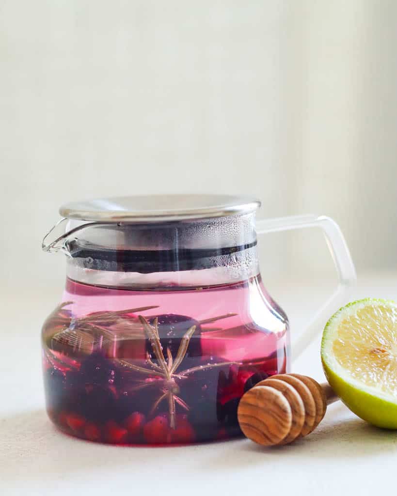 A glass tea infuser filled with blueberries, pomegranate, and rosemary to make rosemary tea the liquid in the infuser is purple due to the blueberries and next to the infuser is a honey stick and lemon