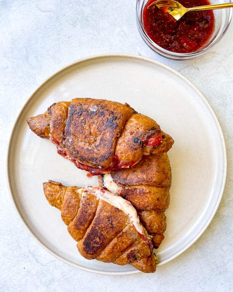 two croissants stuffed with cream cheese and strawberries on a white plate and a small ramekin of homemade strawberry jam in the corner