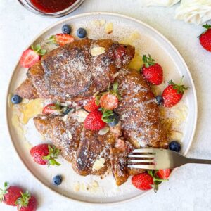 three french toast stuffed croissants on a white plate with strawberries and a fork