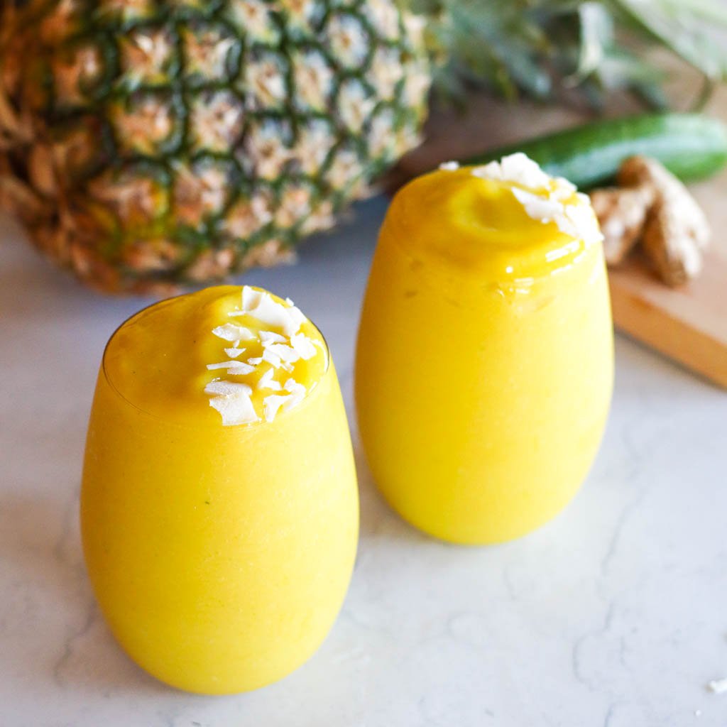 two glasses full of mango pineapple smoothie with ingredients in the background
