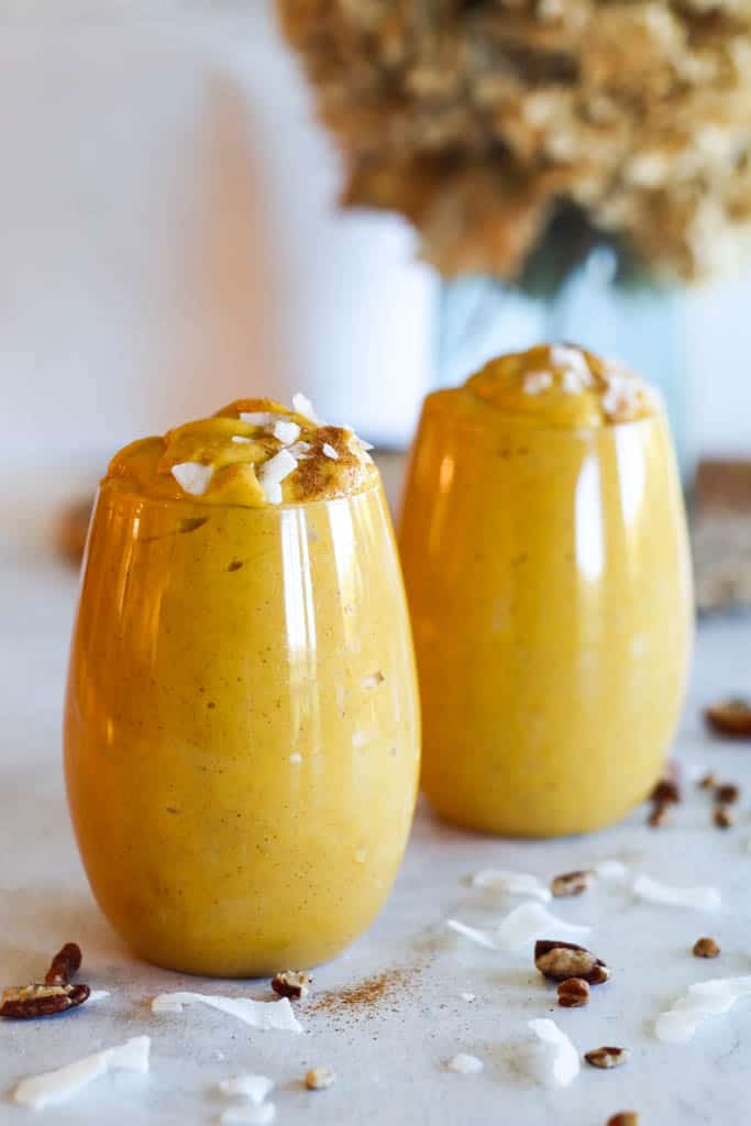 two sweet potato smoothies in glasses and topped with coconut shavings and pecans with beige flowers in the background