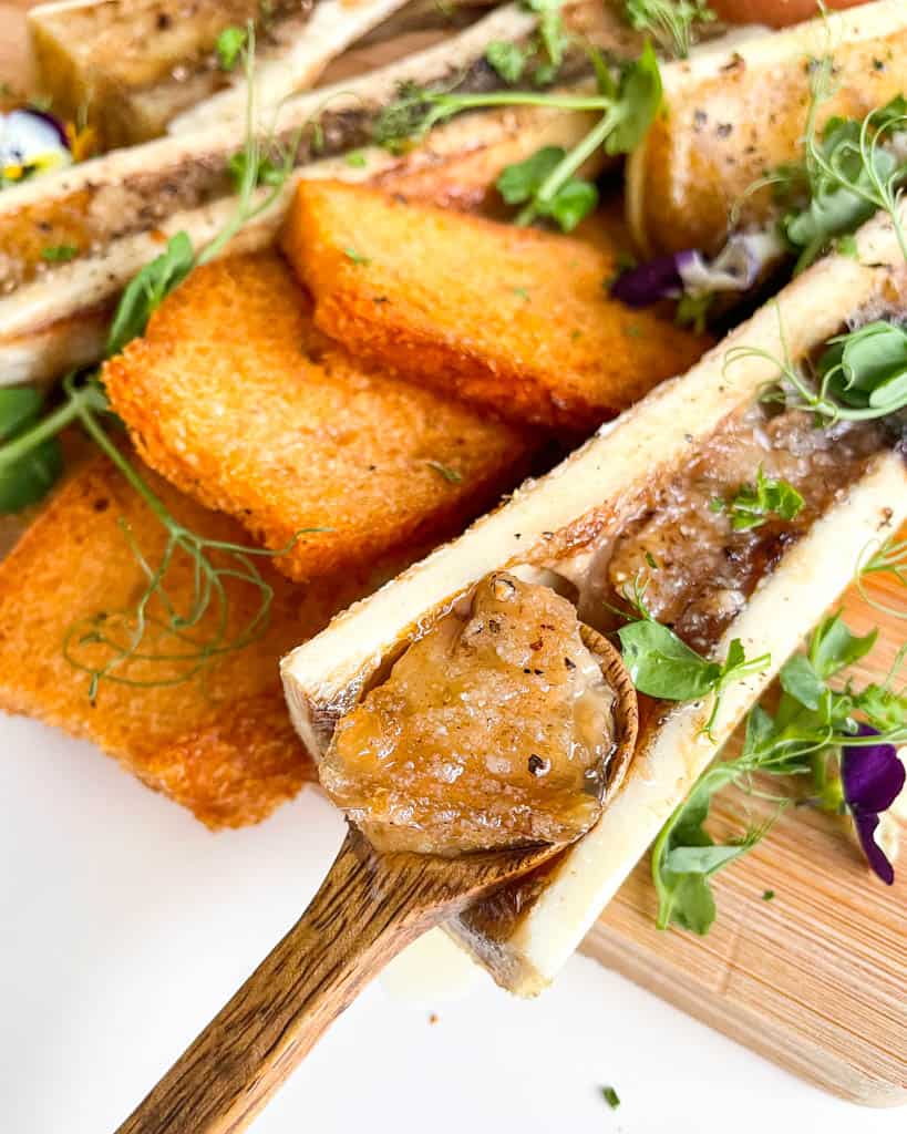 a small wooden spoon with bone marrow on the spoon and placed on a wooden board of bone marrow bones with toasted bread