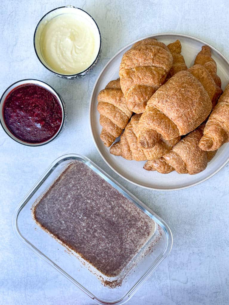 a plate of gluten free croissants a small ramekin of maple cream cheese a small ramekin of strawberry jam and a glass bowl of egg custard to make croissant stuffed french toast