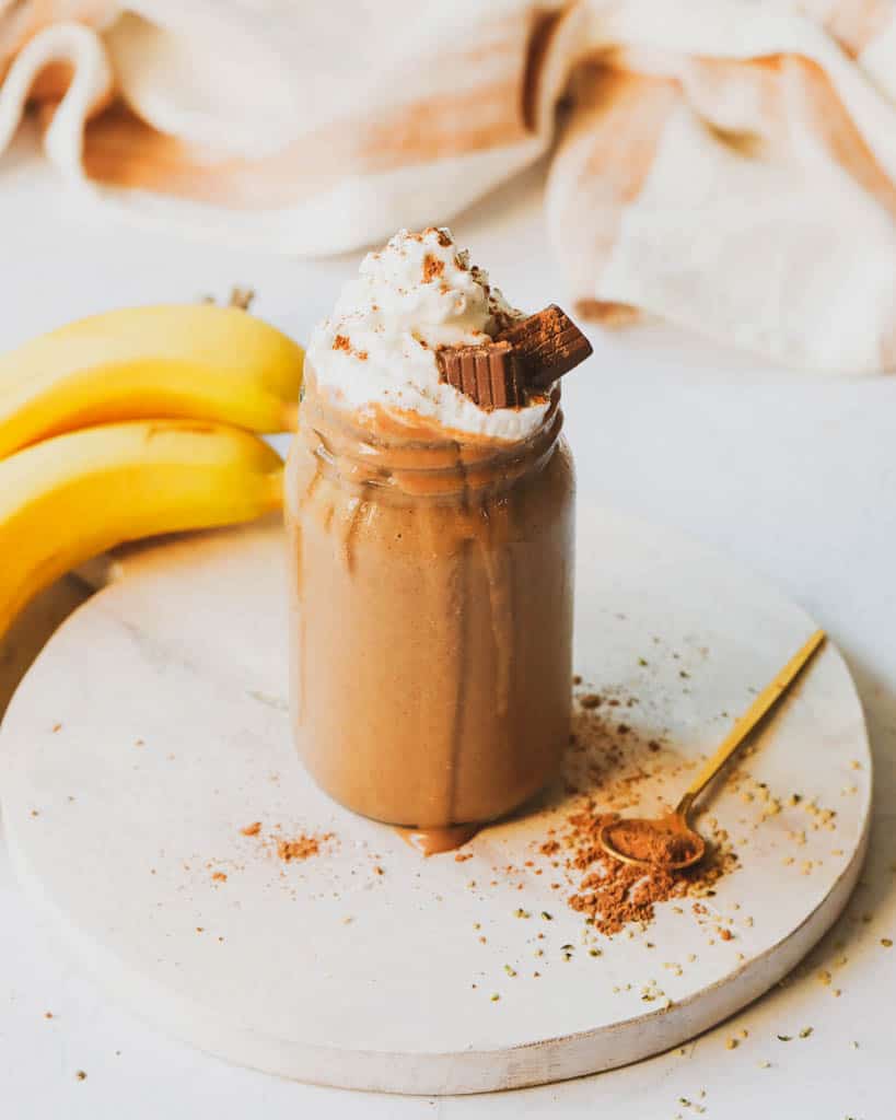 a mason jar full of maca smoothie with bananas in the background and a spoon with maca powder in the forefront