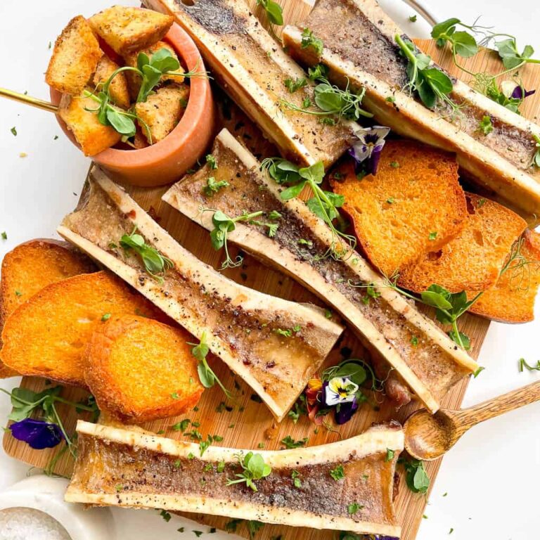 oven roasted bone marrow bones arranged on a wooden board with toasty bread microgreens croutons salt and edible flowers