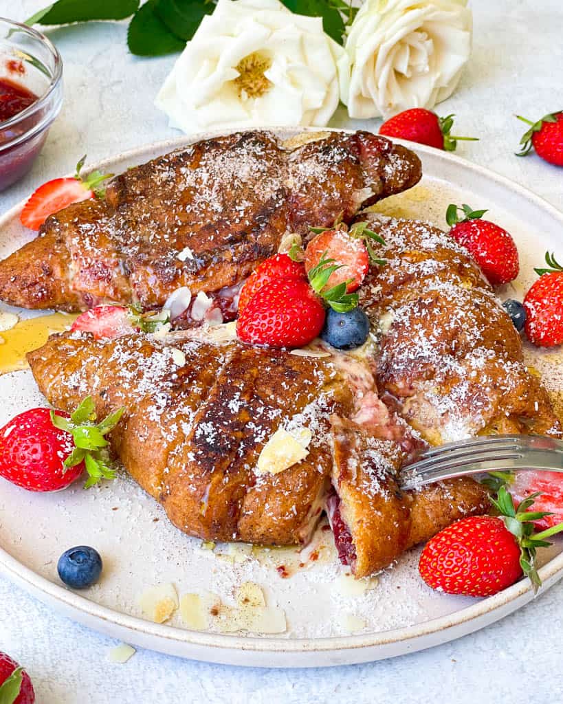 three french toast stuffed croissants on a white plate with strawberries and a fork showing the jammy inside of a slice of the croissant