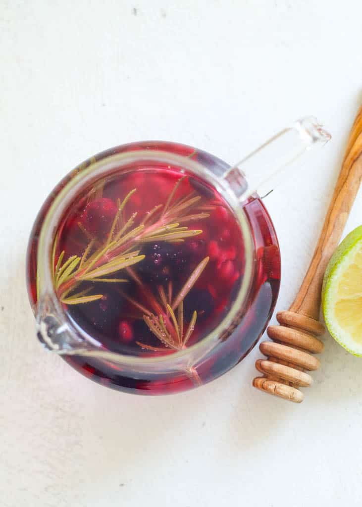 overhead view of A glass tea infuser filled with blueberries, pomegranate, and rosemary to make rosemary tea the liquid in the infuser is purple due to the blueberries and next to the infuser is a honey stick and lemon