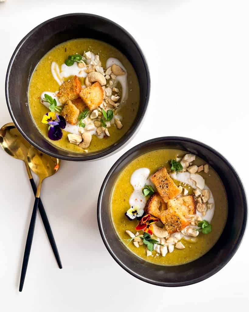 overhead view of two black bowls filled with green soup and topped with chopped cashews and croutons with 2 gold and black spoons next to the bowl