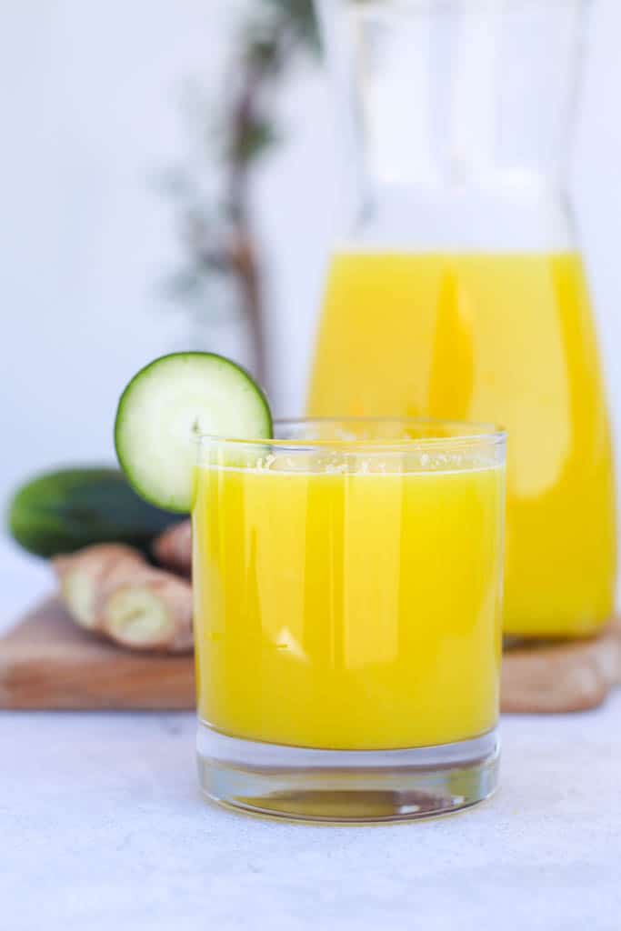 a glass cup full of pineapple and cucumber juice garnished with a slice of cucumber with a carafe filled with more juice in the background