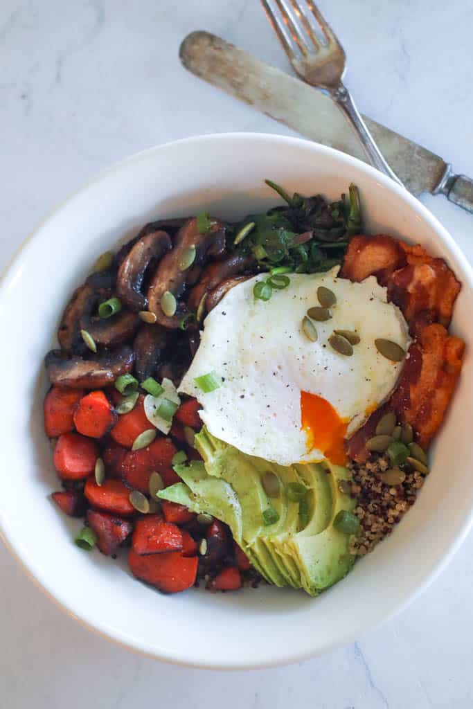 overhead view of a quinoa breakfast bowl with egg, avocado, bacon, mushrooms, and carrots with a fork and knife beside it