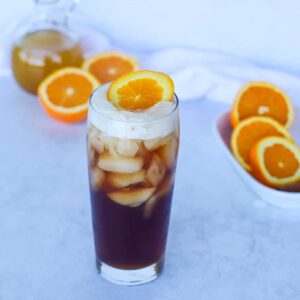 a glass full of cold brew tonic garnished with an orange slice and more sliced oranges in the background