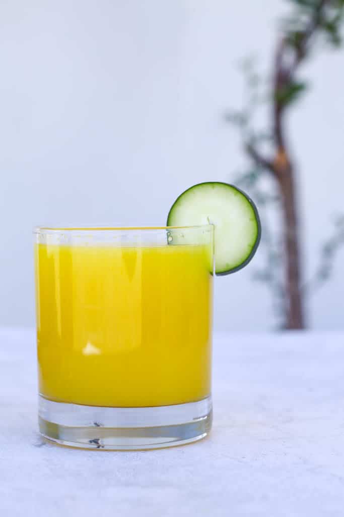 a glass cup full of pineapple and cucumber juice garnished with a slice of cucumber