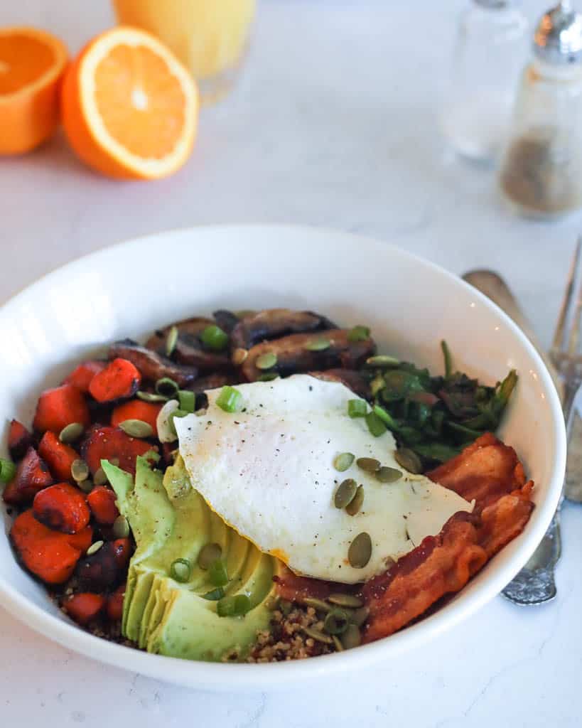 a quinoa breakfast bowl with egg, avocado, bacon, mushrooms, and carrots with halved oranges in the background