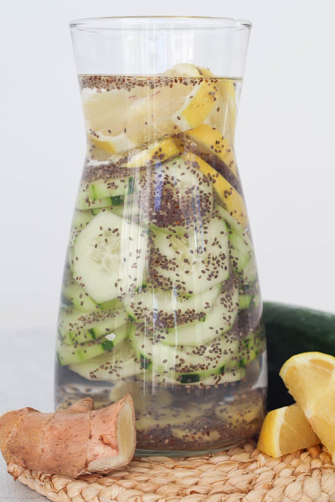 a carafe full of cucumber lemon ginger water with ginger and sliced lemon beside it