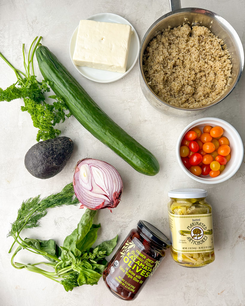 ingredients to make greek quinoa salad shown is a large block of feta a pan of cooked quinoa red onion a small bowl of cherry tomatoes a jar of marinated artichoke a jar of kalamata olives a large english cucumber and fresh herbs