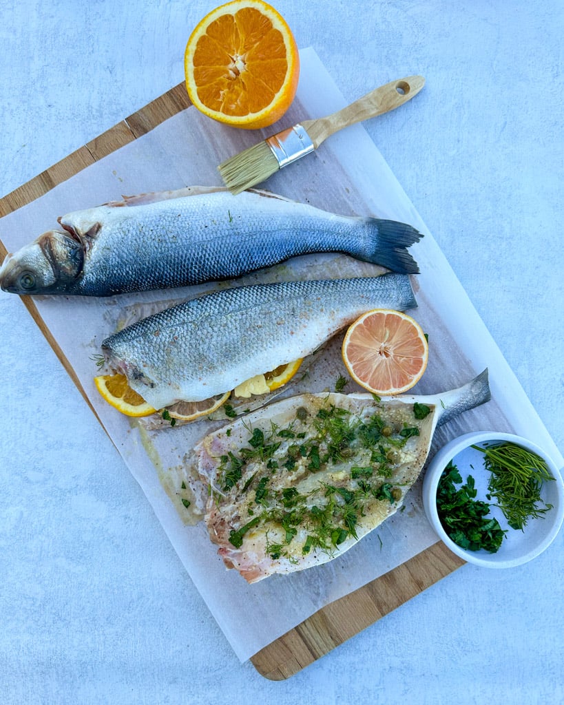 three whole branzino stuffed with lemon orange herbs and garlic on a parchment lined cutting board with one piece butterflied open to show herbs and seasoned flesh