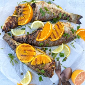 three pieces of whole branzino grilled stuffed with lemon herbs and garlic with sliced lemon and orange on top of the branzino