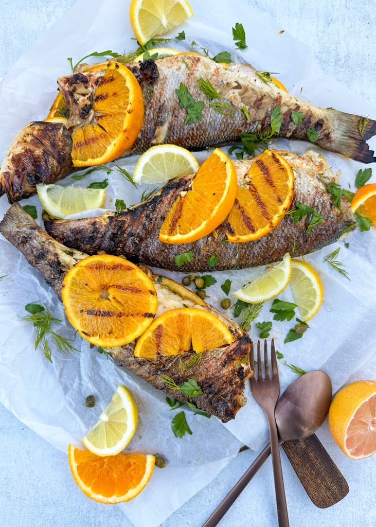 three pieces of whole branzino grilled stuffed with lemon herbs and garlic with sliced lemon and orange on top of the branzino 