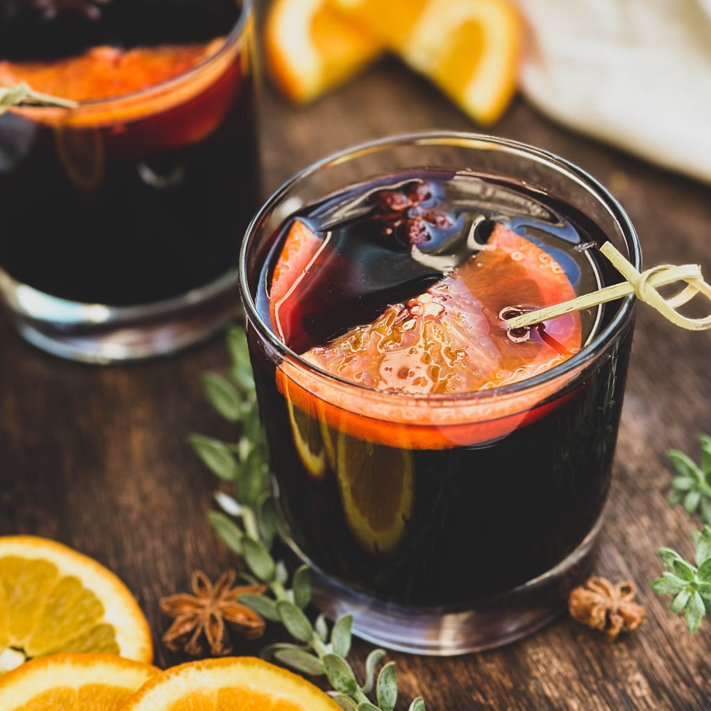 two glasses of mulled wine with orange slices on top and the glasses are placed on a wooden board with greenery orange slices and whole spices around the glasses