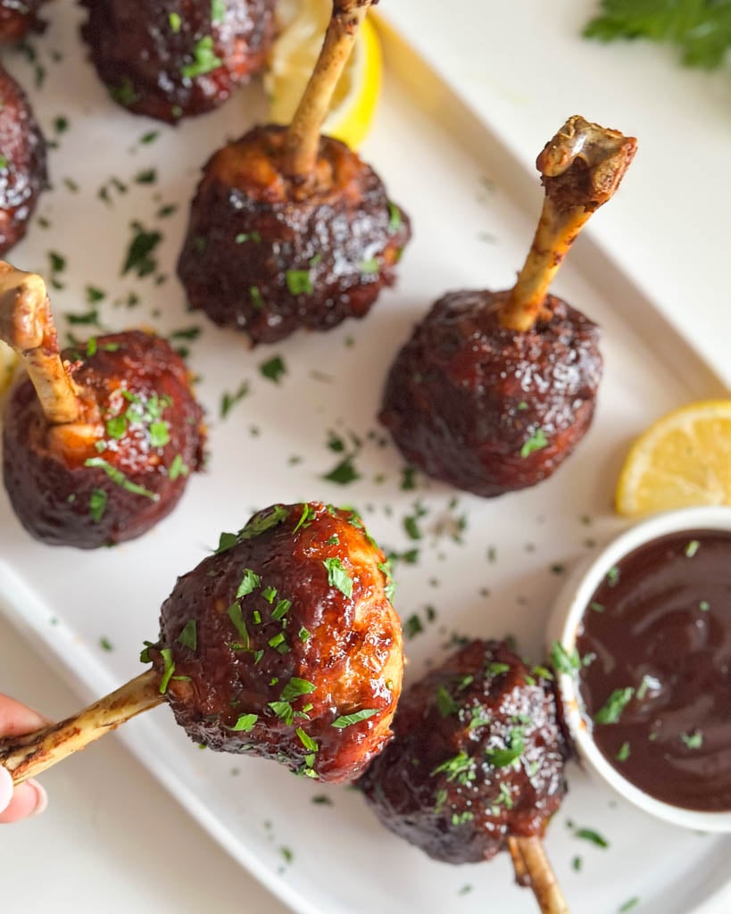 holding chicken lollipop drumstick close to camera with a plate of four lollipop drumsticks in the background and a small ramekin of barbecue sauce on the plate