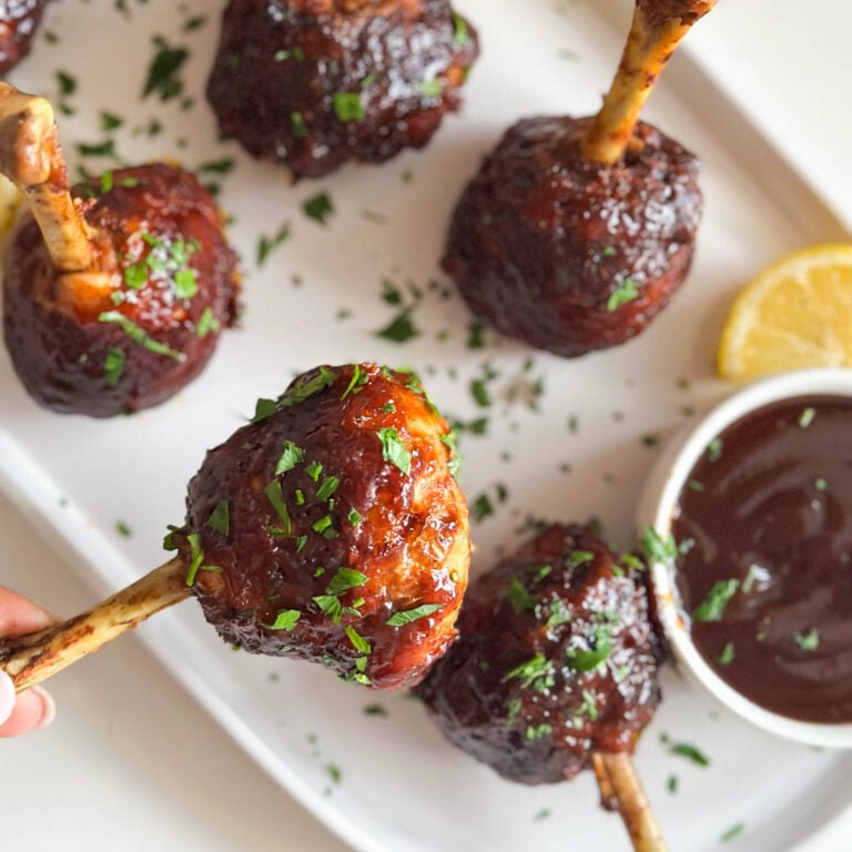 holding chicken lollipop drumstick close to camera with a plate of four lollipop drumsticks in the background and a small ramekin of barbecue sauce on the plate