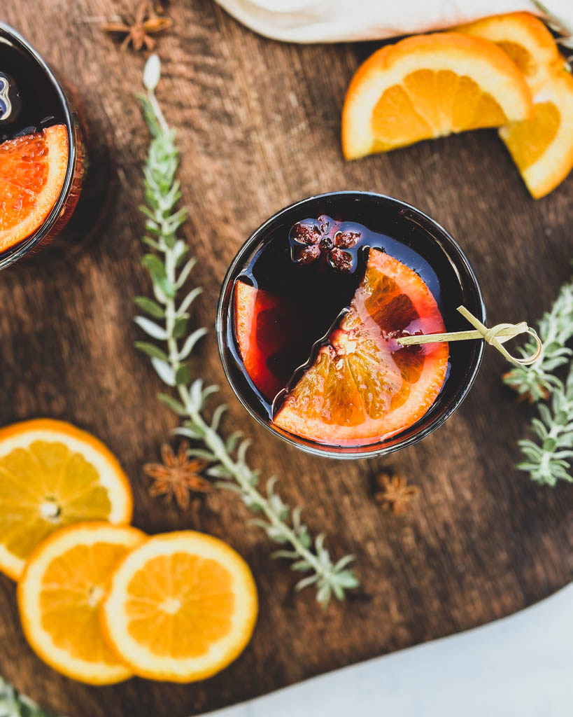 a glass of spiced wine with orange slices on top and the glass is placed on a wooden board with greenery orange slices and whole spices around the glasses