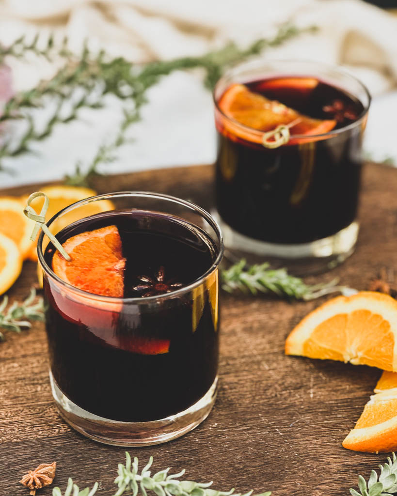two glasses of mulled wine with orange slices on top glasses are places on a wooden board with greenery orange slices and whole spices around the glasses