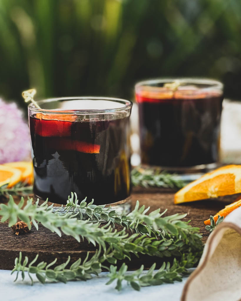 two glasses of mulled wine with orange slices on top and the glasses are placed on a wooden board with greenery orange slices and whole spices around the glasses