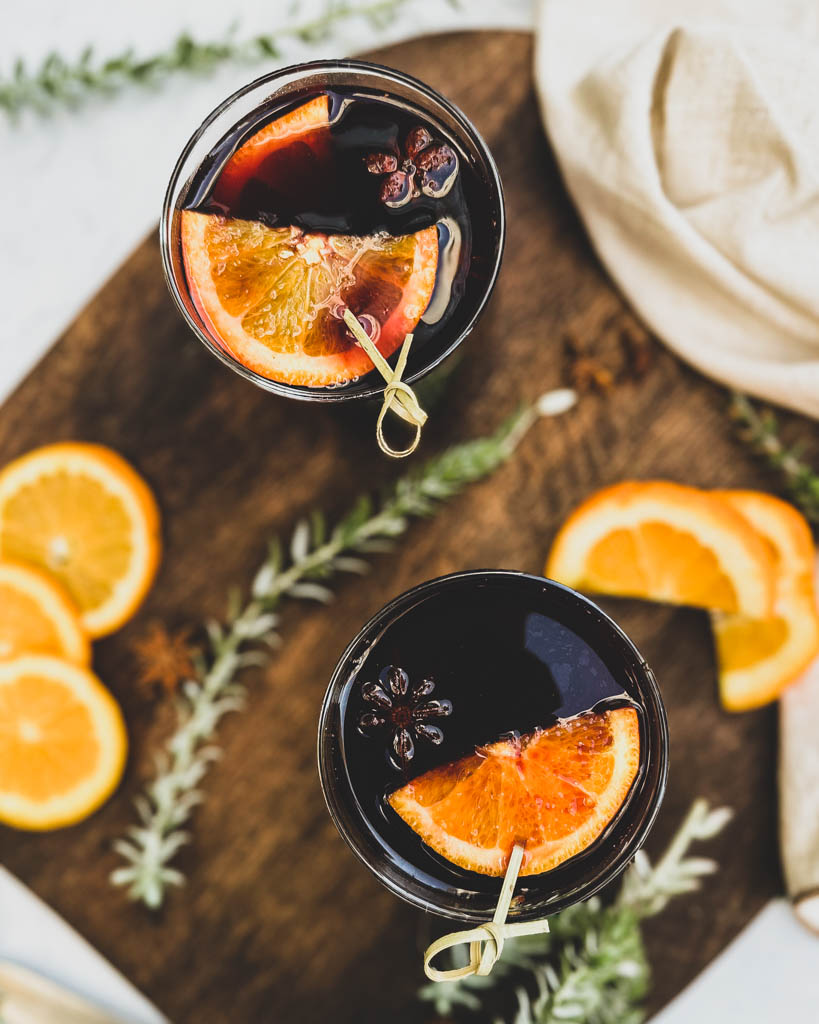 overhead image of two glasses of mulled wine with orange slices on top and the glasses are placed on a wooden board with greenery orange slices and whole spices around the glasses