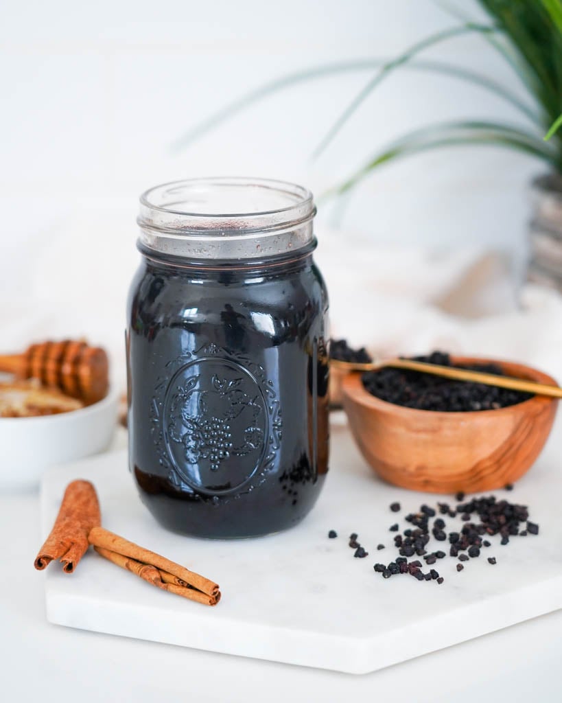 a jar of elderberry syrup surrounded by honey, cinnamon sticks, and a small bowl of dried elderberries