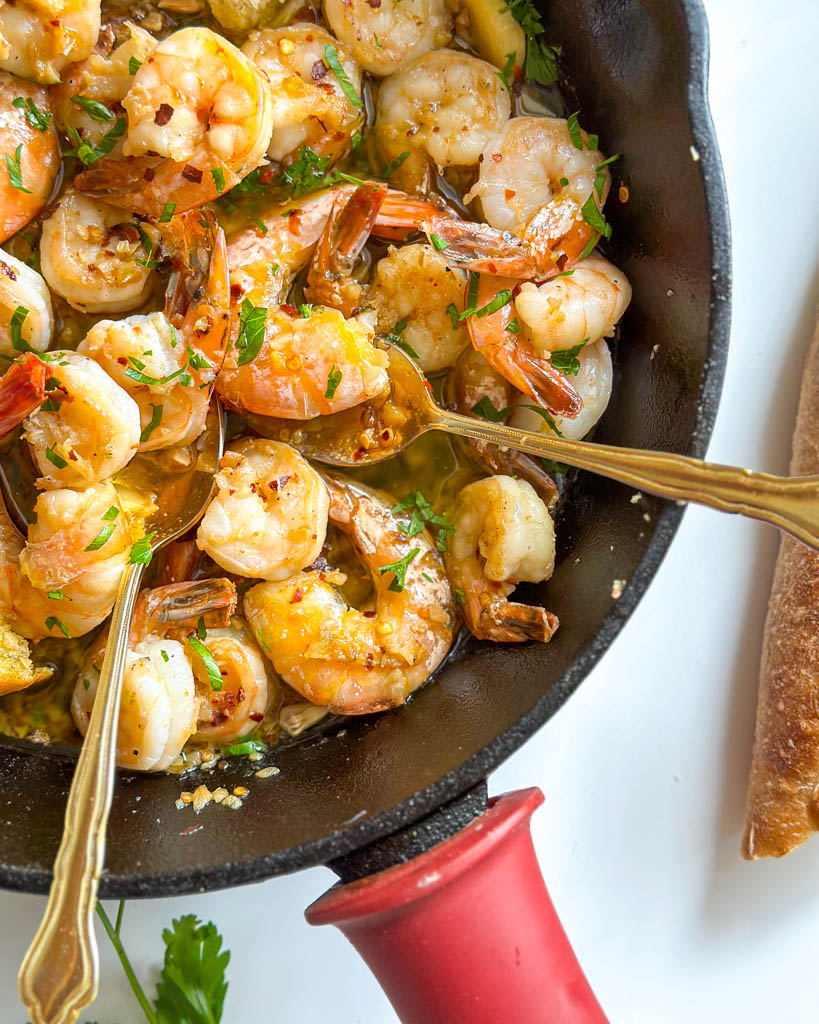 gambas al ajillo also known as spanish garlic shrimp shown in a cast iron with two gold spoons in it