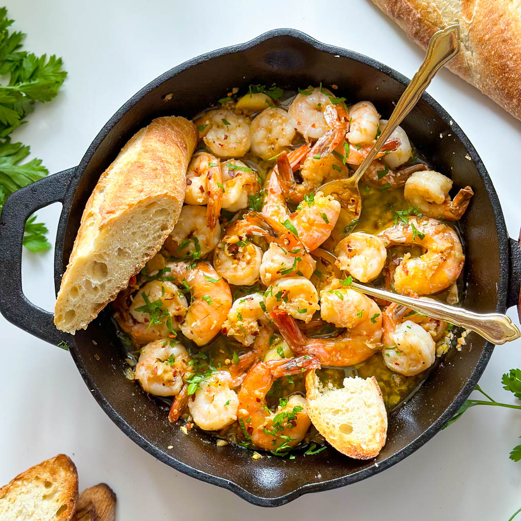 gambas al ajillo also known as spanish garlic shrimp shown in a cast iron with two gold spoons in it and parsley bread loaf and small bowl of red crushed peppers on the side of the cast iron