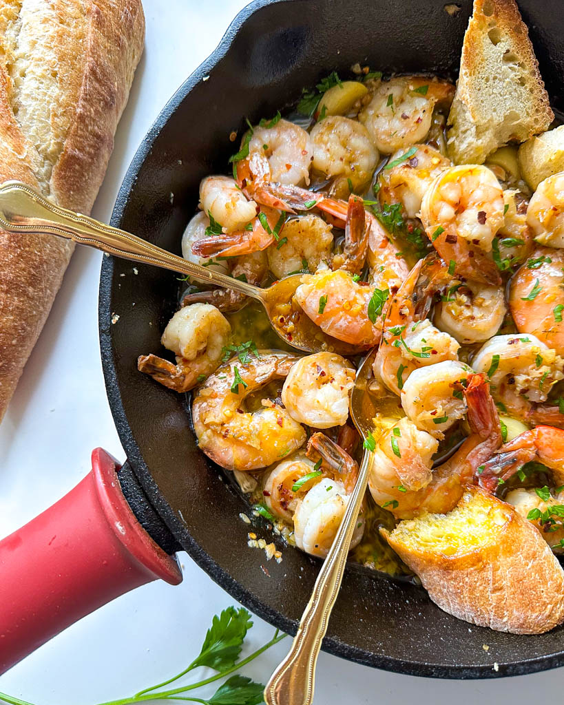 gambas al ajillo also known as spanish garlic shrimp shown in a cast iron with two gold spoons in it and parsley bread loaf on the side of the cast iron