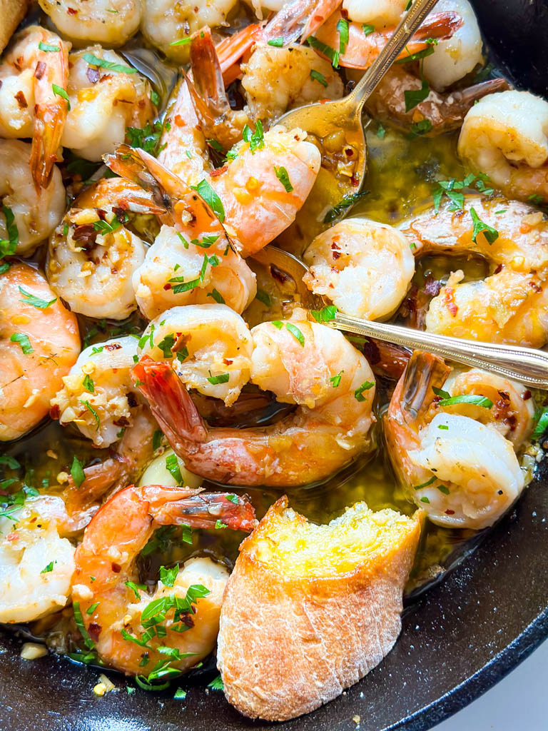 gambas al ajillo also known as spanish garlic shrimp shown in a cast iron with two gold spoons in it
