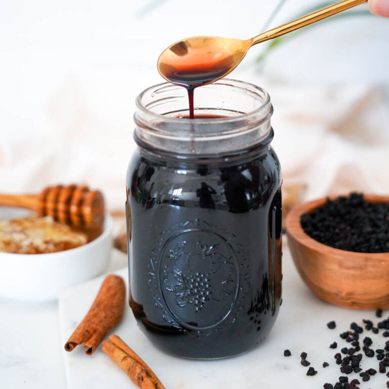 a spoon containing elderberry syrup suspended over a jar of more with honey cinnamon sticks and a small bowl of dried elderberries surrounding it