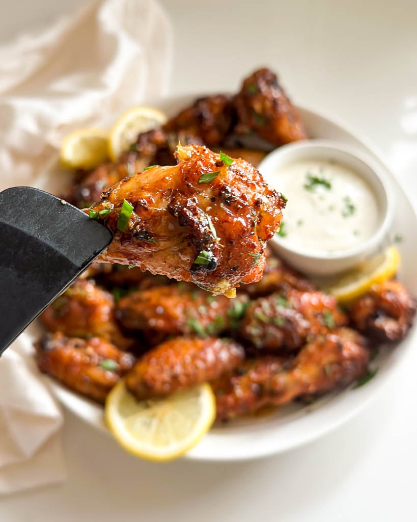 a tong holding a single honey lemon pepper wing over a plate of more wings and ranch dipping sauce
