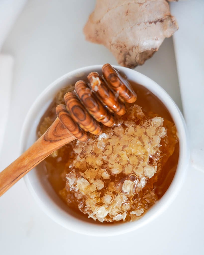 a honey dipper hovering over a bowl of honey with ginger in the background