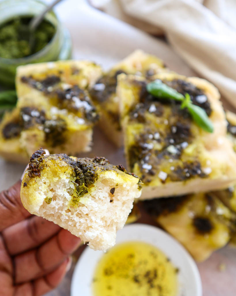 a hand holding a chunk of Gluten Free Focaccia bead with a saucer of olive oil and a jar of pesto in the background