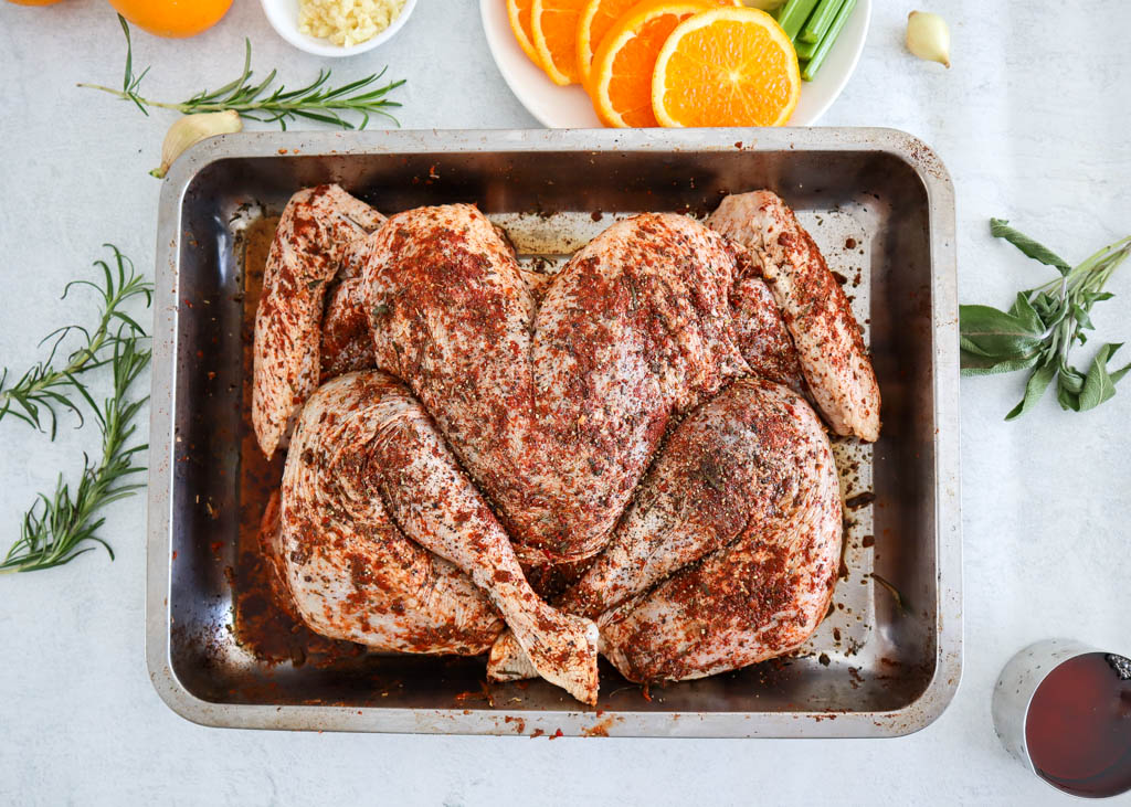 dry brine seasoning rubbed on the front side of a butterflied turkey placed in a roasting tray with sliced oranges and fresh herbs on the side of the tray