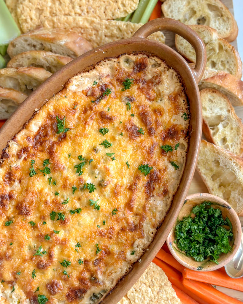 a large dish of hot crab dip served on a board with celery parmesan crisp carrots and sliced bread
